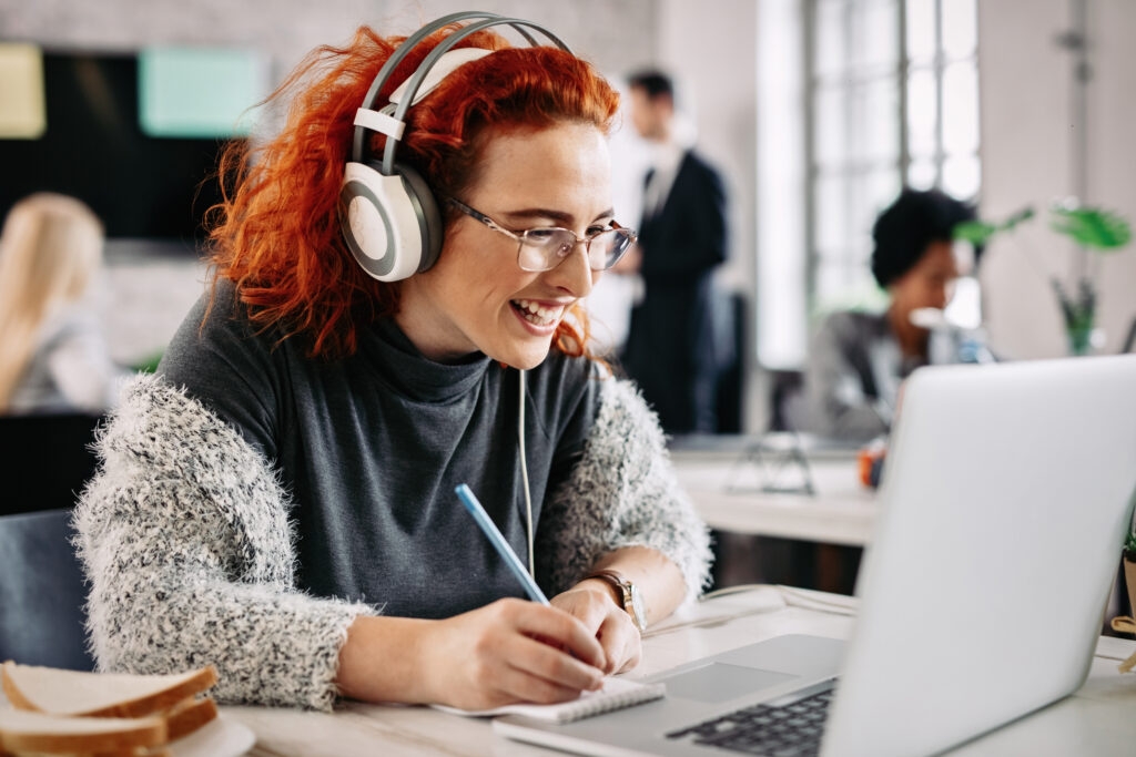 Cheerful female entrepreneur working on a computer and taking notes in her notepad while listening music on headphones in the office. There are people in the background.